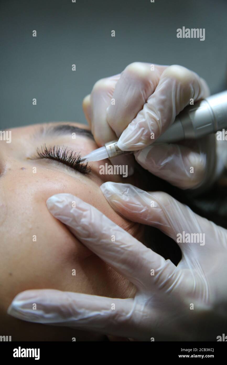 woman lying on couch at beauty salon with closed eyes and enjoying when cosmetologist making eyeliner and eyebrow permanent make up. Professional wear Stock Photo