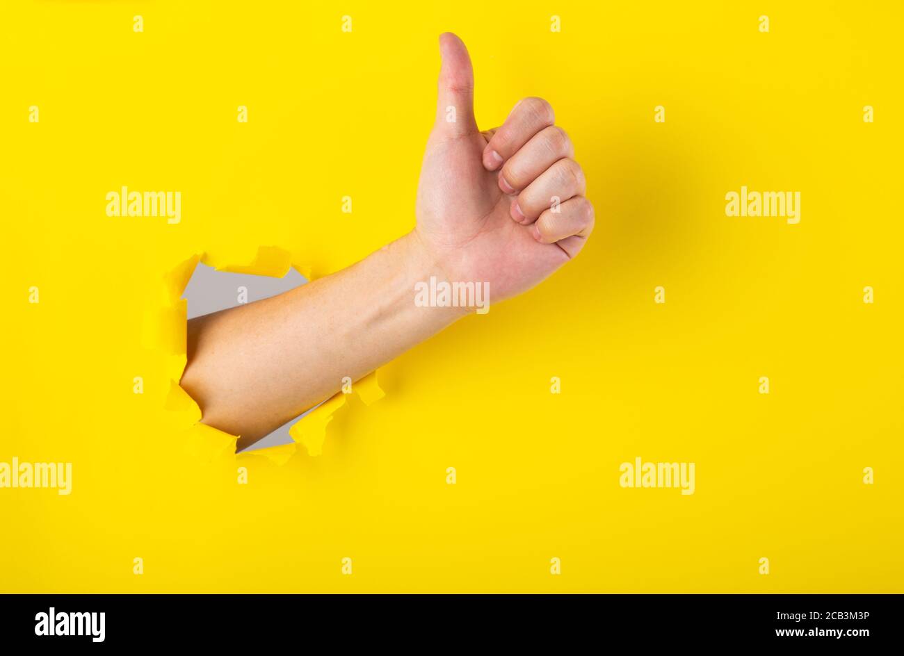 Hand showing a thumb up sign through a ripped hole in a yellow paper ...