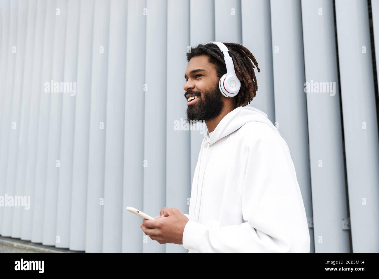 Image of a handsome pleased smiling young man walking outdoors while listening music by headphones and using mobile phone Stock Photo