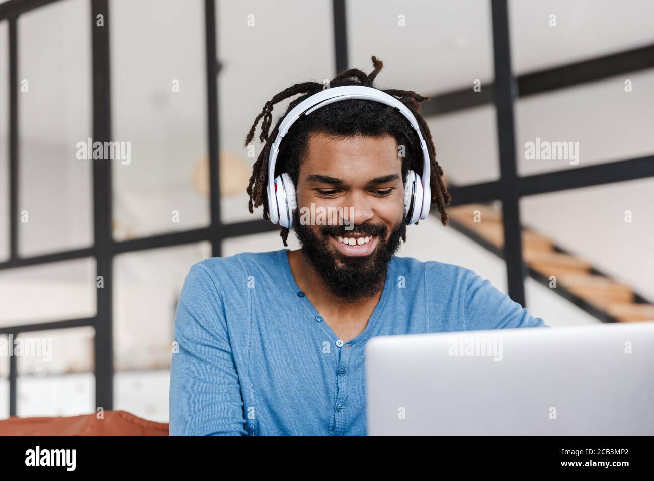 Handsome smiling young african man sitting on a leather couch at home, using laptop computer, wearing wireless headphones Stock Photo