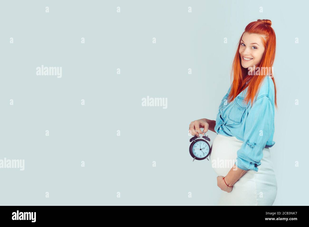 Clock is ticking, expectation concept. Side view of smiling young woman with baby in tummy holding alarm clock waiting for birth and smiling at camera Stock Photo