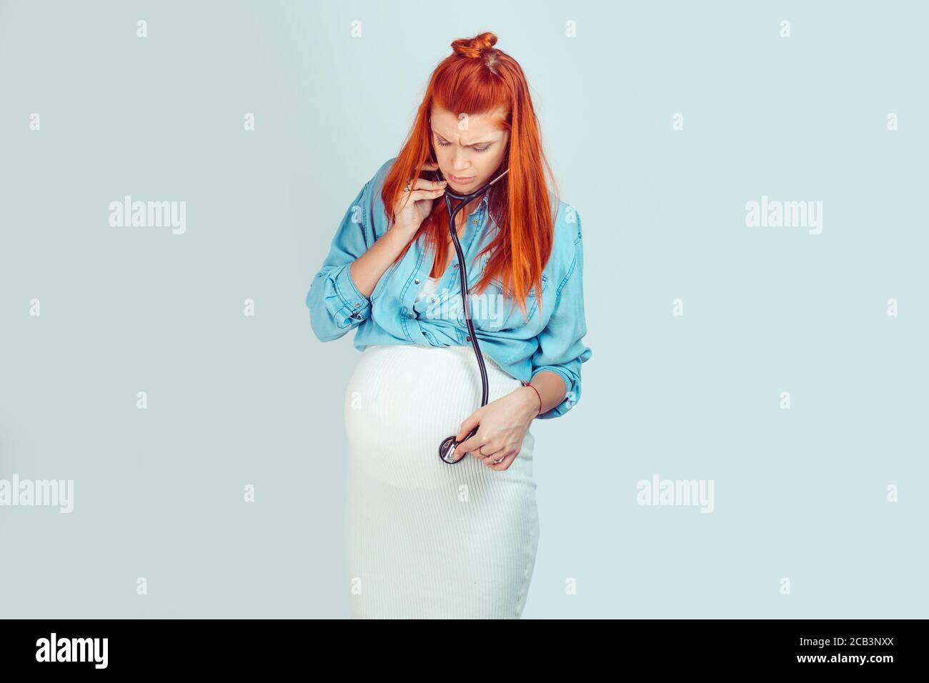 Young ginger pregnant woman using stethoscope and listening to baby with worries isolated on