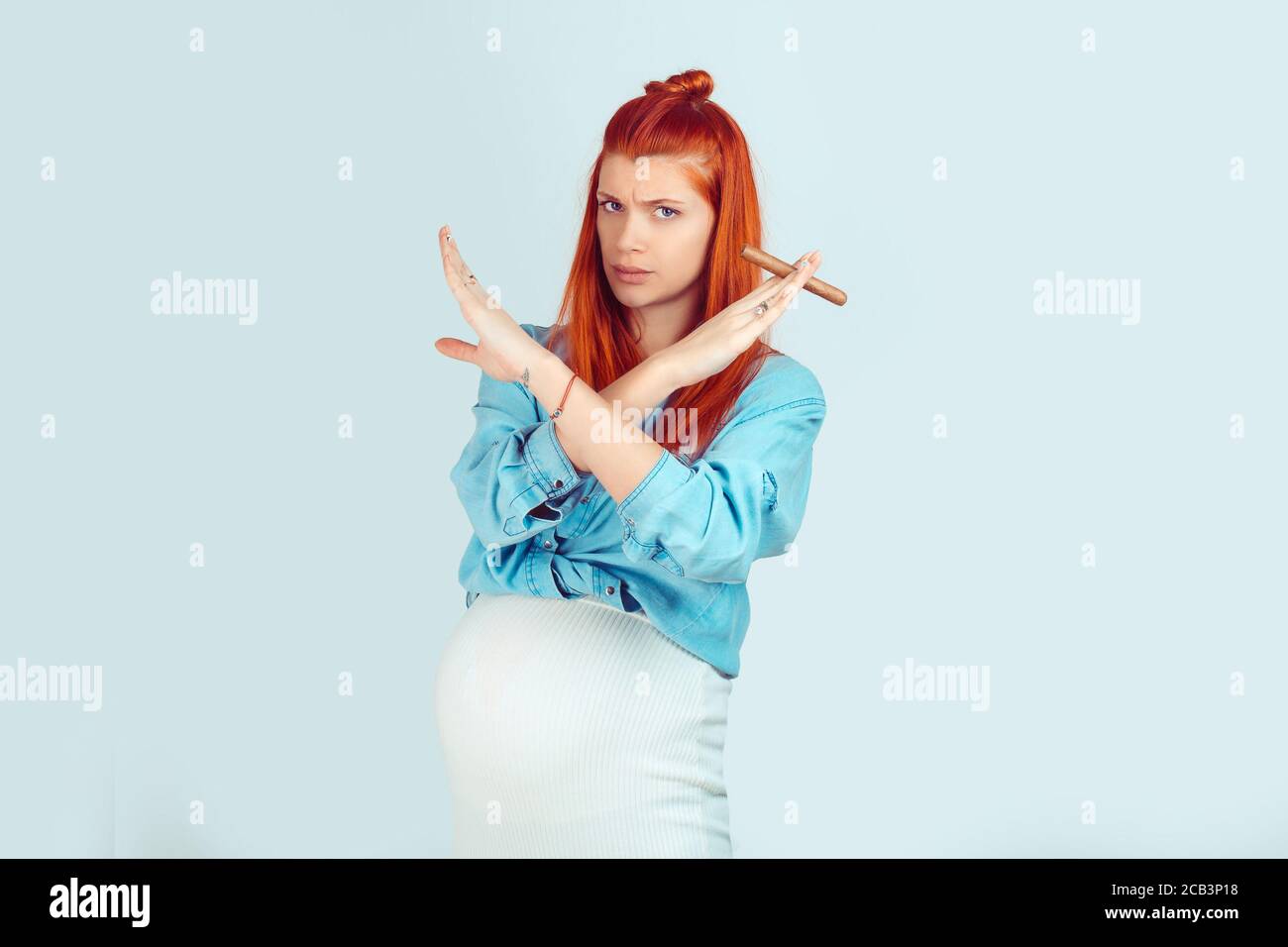 Charming ginger girl with baby in belly holding hands crossed with cigar looking extremely against smoking isolated on blue backdrop. Mixed race model Stock Photo