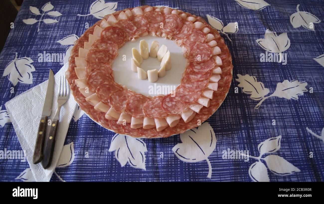 White plate decorated with salami, ham and heart of palm in the center on a background with a blue and white tablecloth, with a knife and fork, a dish Stock Photo