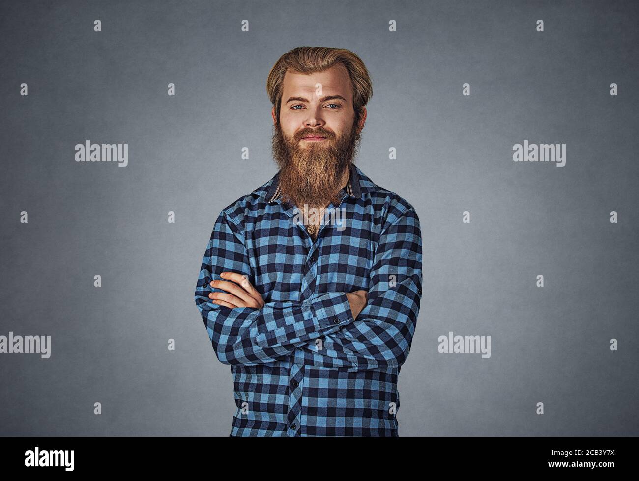 Young bearded man looking at you camera hands folded. Hipster male with beard in blue plaid checkered shirt  Isolated on gray studio Background. Negat Stock Photo