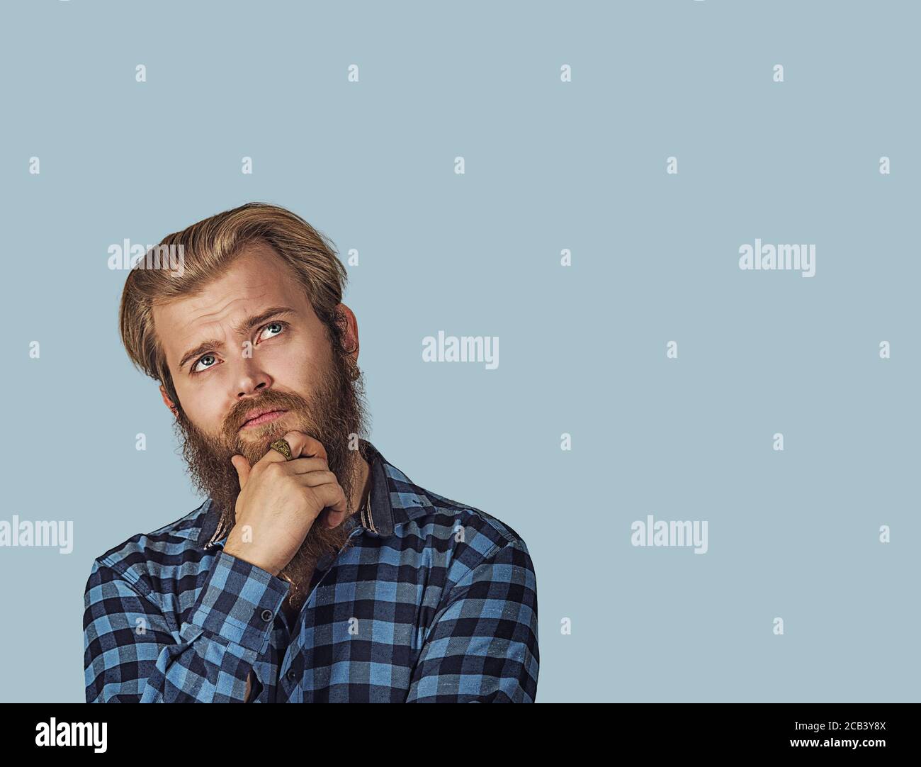 Confused skeptical man thinking looking up with copy space above head. Hipster male with beard in blue plaid checkered shirt  Isolated on blue studio Stock Photo