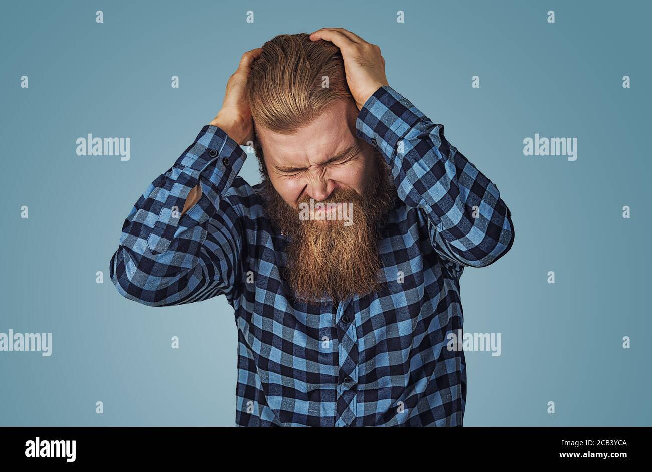 Depressed man having a very strong headache. Hipster male with beard in blue plaid checkered shirt  Isolated on blue studio Background. Negative face Stock Photo
