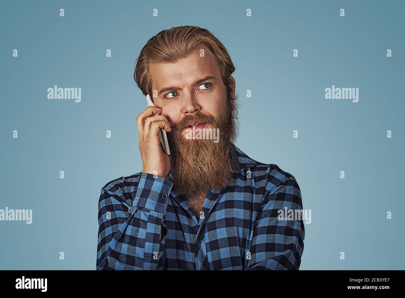 Cool serious man with beard talking on phone and looking up to side. Casual young man talking on smartphone. Hipster male with beard in blue plaid che Stock Photo