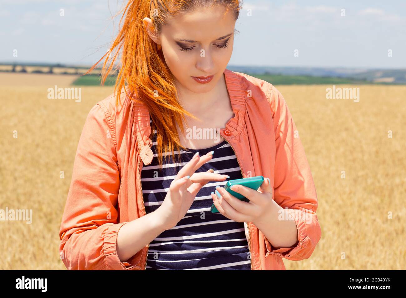 No coverage. Pretty redhead girl woman texting, checking, sending a message looking for coverage on her mobile phone isolated yellow orange wheat fiel Stock Photo