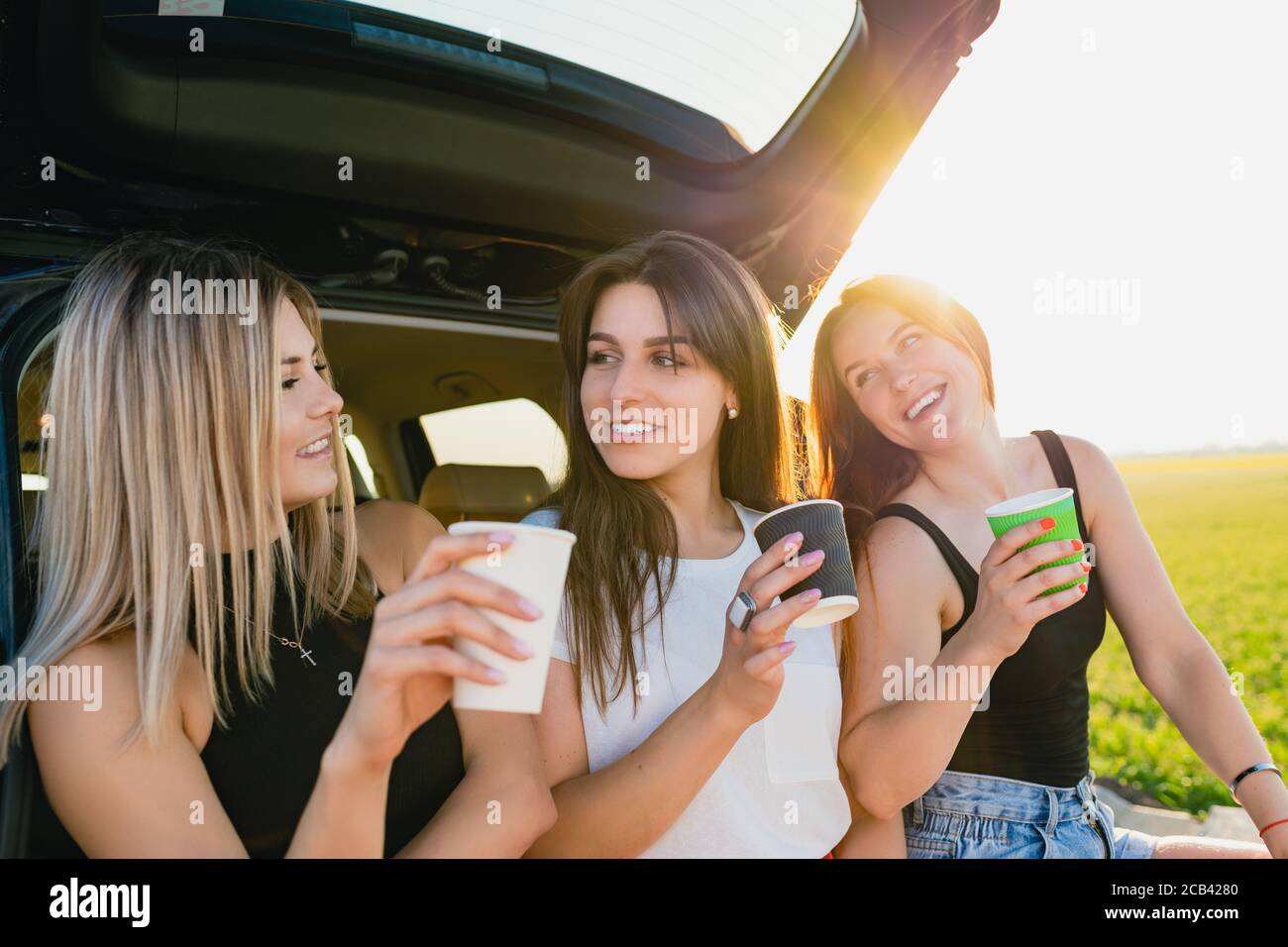 Happy road trip of three smiling female travelers, relax at vehicle back with coffee cups take away at sunlight. The girls are drinking coffee. Stock Photo