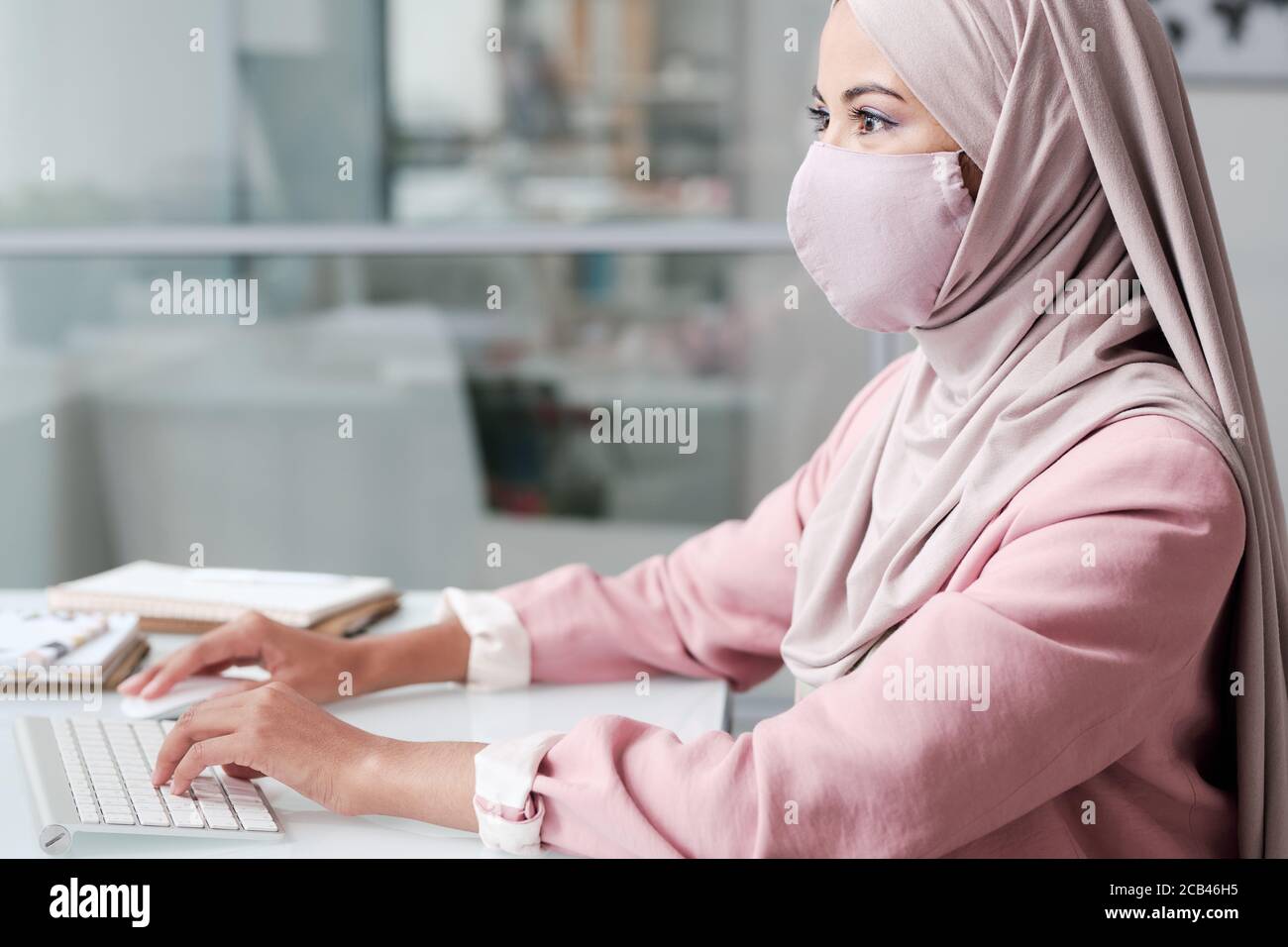 Side view of busy young Muslim woman in facial mask working in office ...