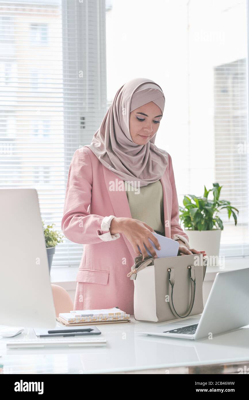 Attractive middle-eastern businesswoman in pink hijab standing at desk and packing phone in bag while going to leave office Stock Photo