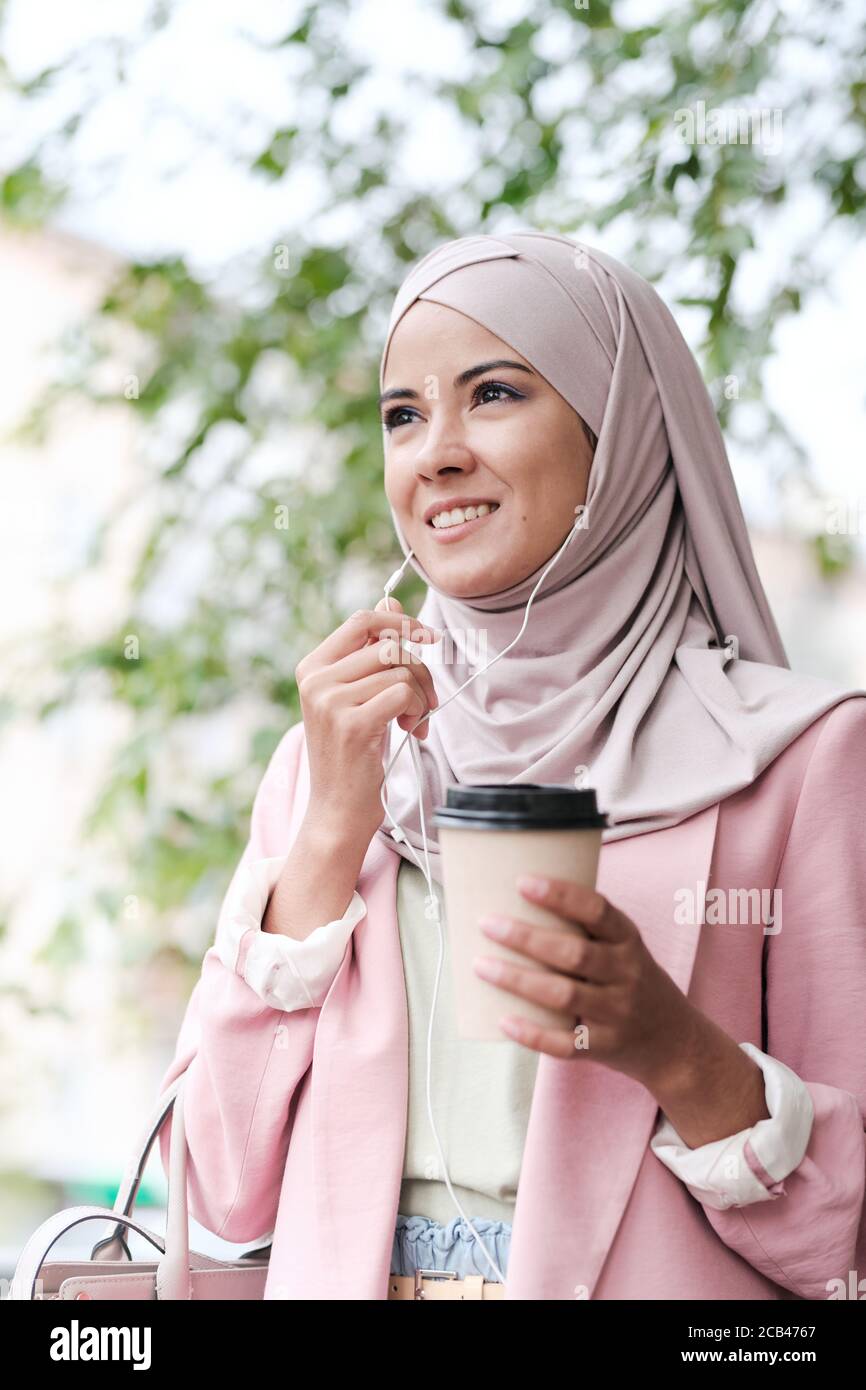 Below view of smiling confident young Arabian businesswoman in hijab drinking coffee and using earpieces while answering phone on move Stock Photo