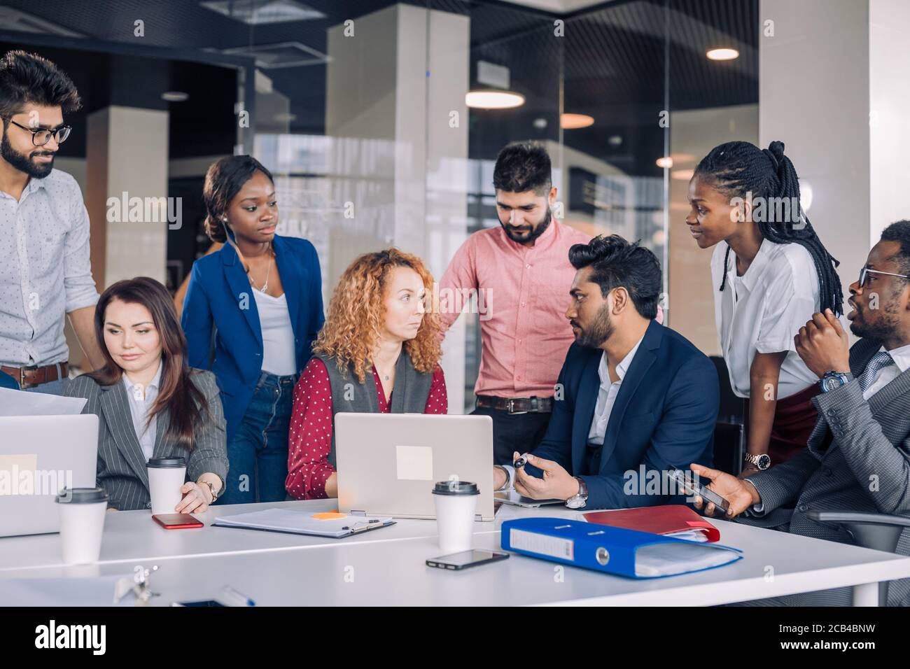 Creative multiracial business team discussing project, standing around workplace, while two co-workers with portable computers looking for needed info Stock Photo