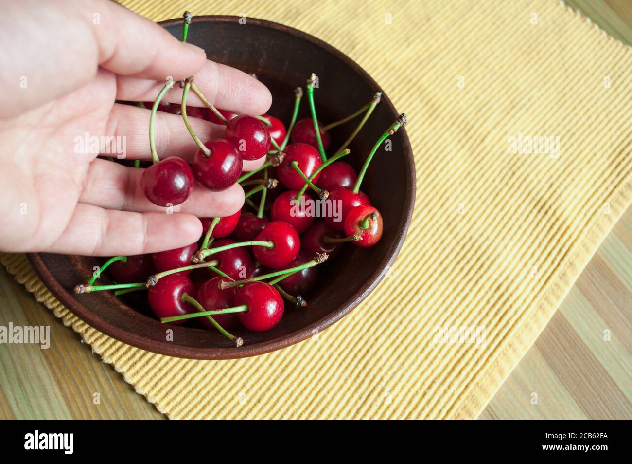 A woman aged 30-40 holds three ripe cherries over a plate of cherries. The photo shows part of the palm and a plate with cherries Stock Photo