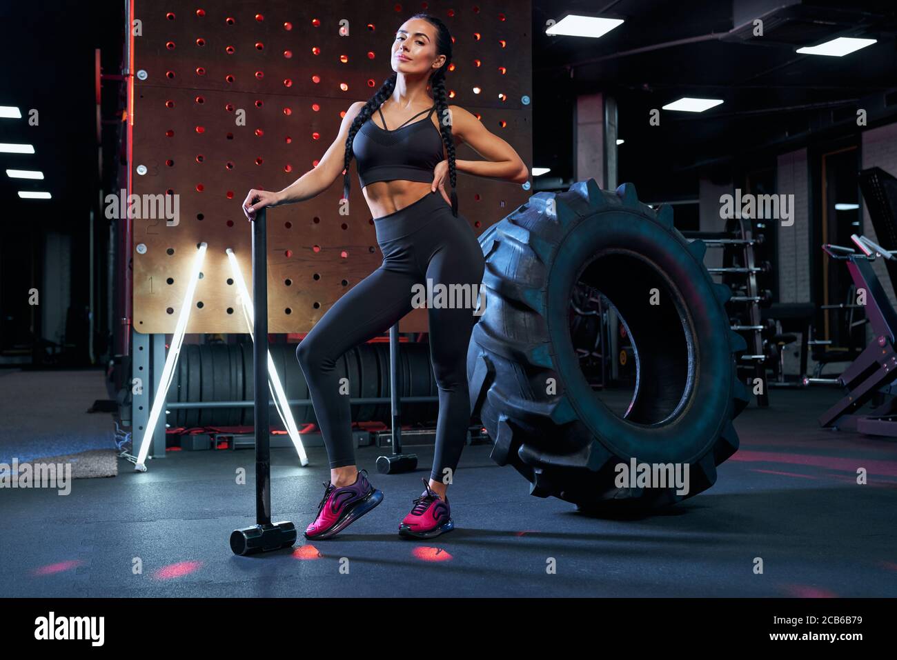 Front view of stunning muscular woman holding big sledgehammer near tire  wheel, looking at camera. Portrait of female bodybuilder in sportswear  posing in gym, having rest after hard training Stock Photo -