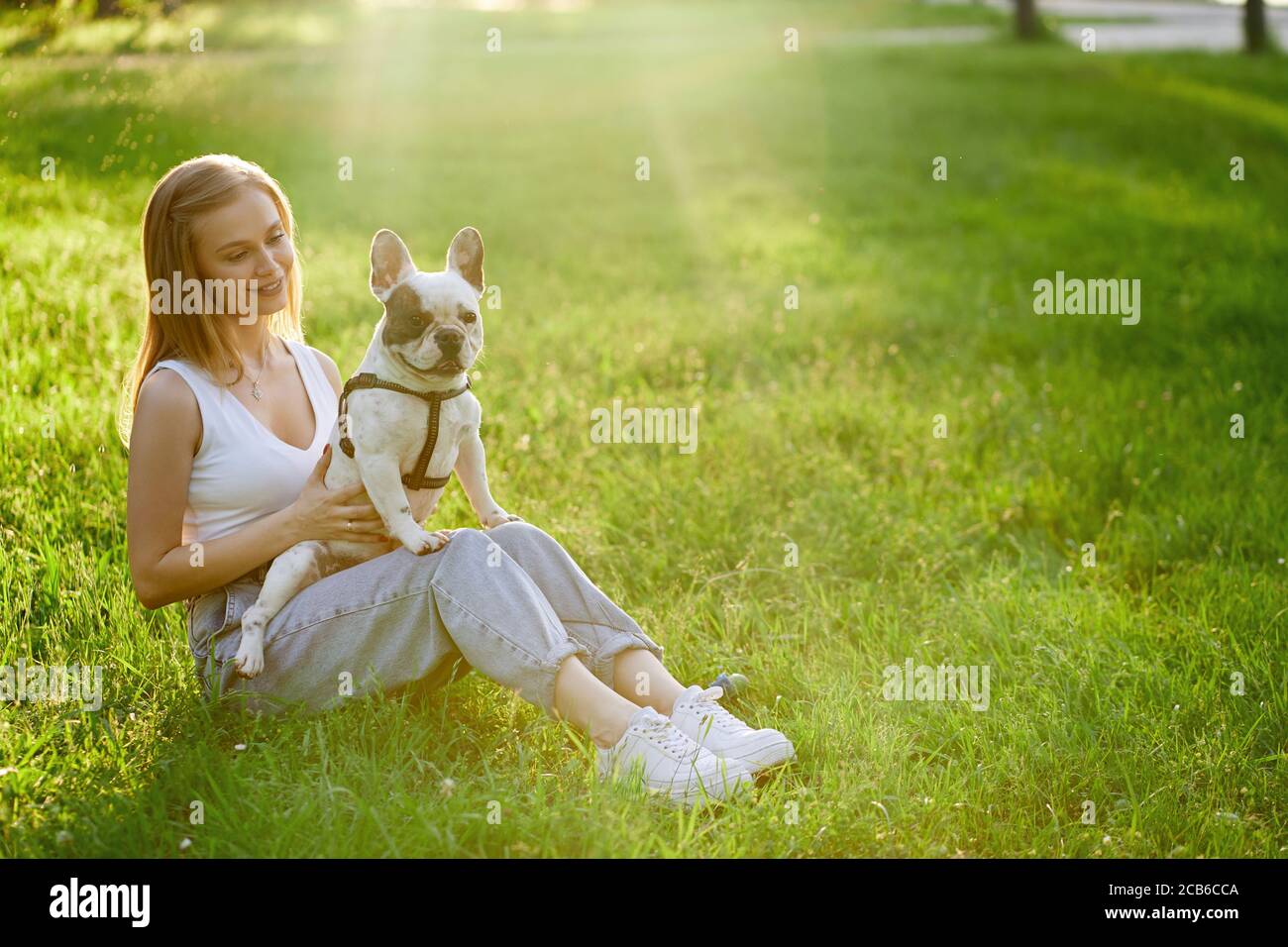 Top view of young happy woman sitting on grass with lovely french bulldog. Gorgeous caucasian smiling girl enjoying summer sunset, holding dog on knees in city park. Human and animal friendship. Stock Photo