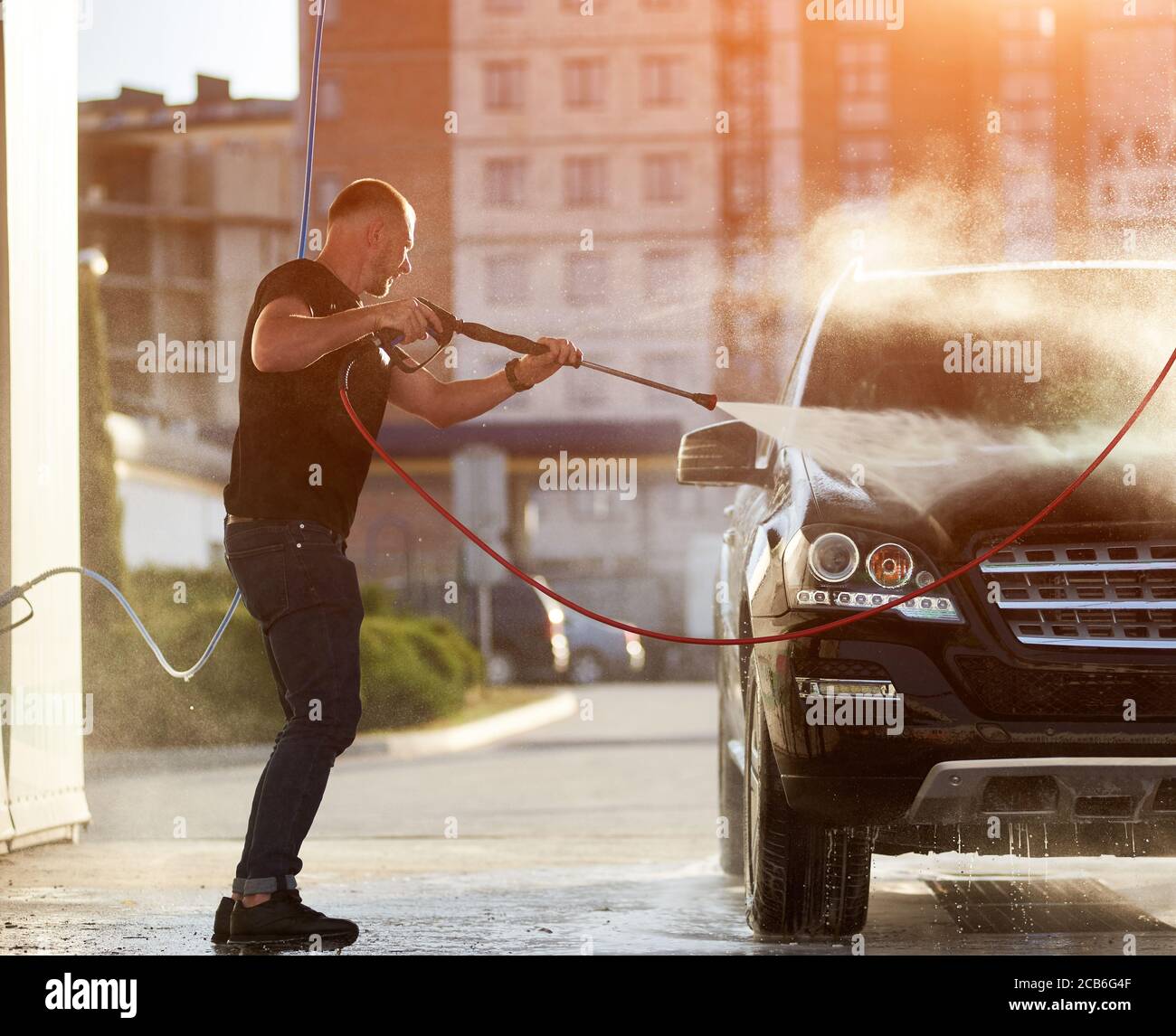 Side view of a sporty man cleaning his black car outdoors, water is splashing over the top of the car shining on the sun, residential building is on the background Stock Photo