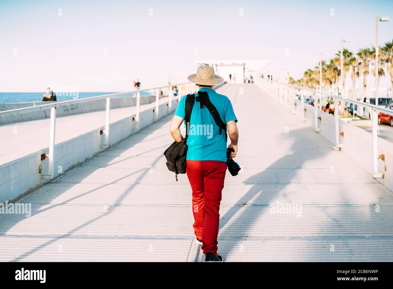 Adult tourist with straw hat and camera hanging from his shoulder walks backwards along a footbridge by the sea. Solo traveler Stock Photo