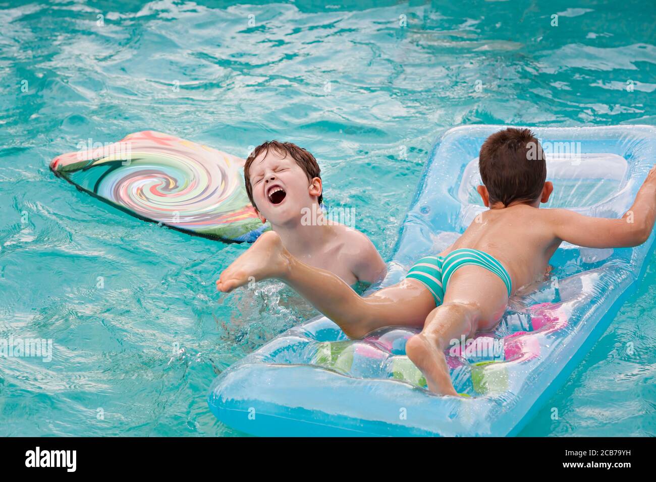 Happy kids playing in the swimming pool with a water mattress. Having fun in the water pool outside on summer day. Boy is having Muscle Cramp While Swimming Stock Photo