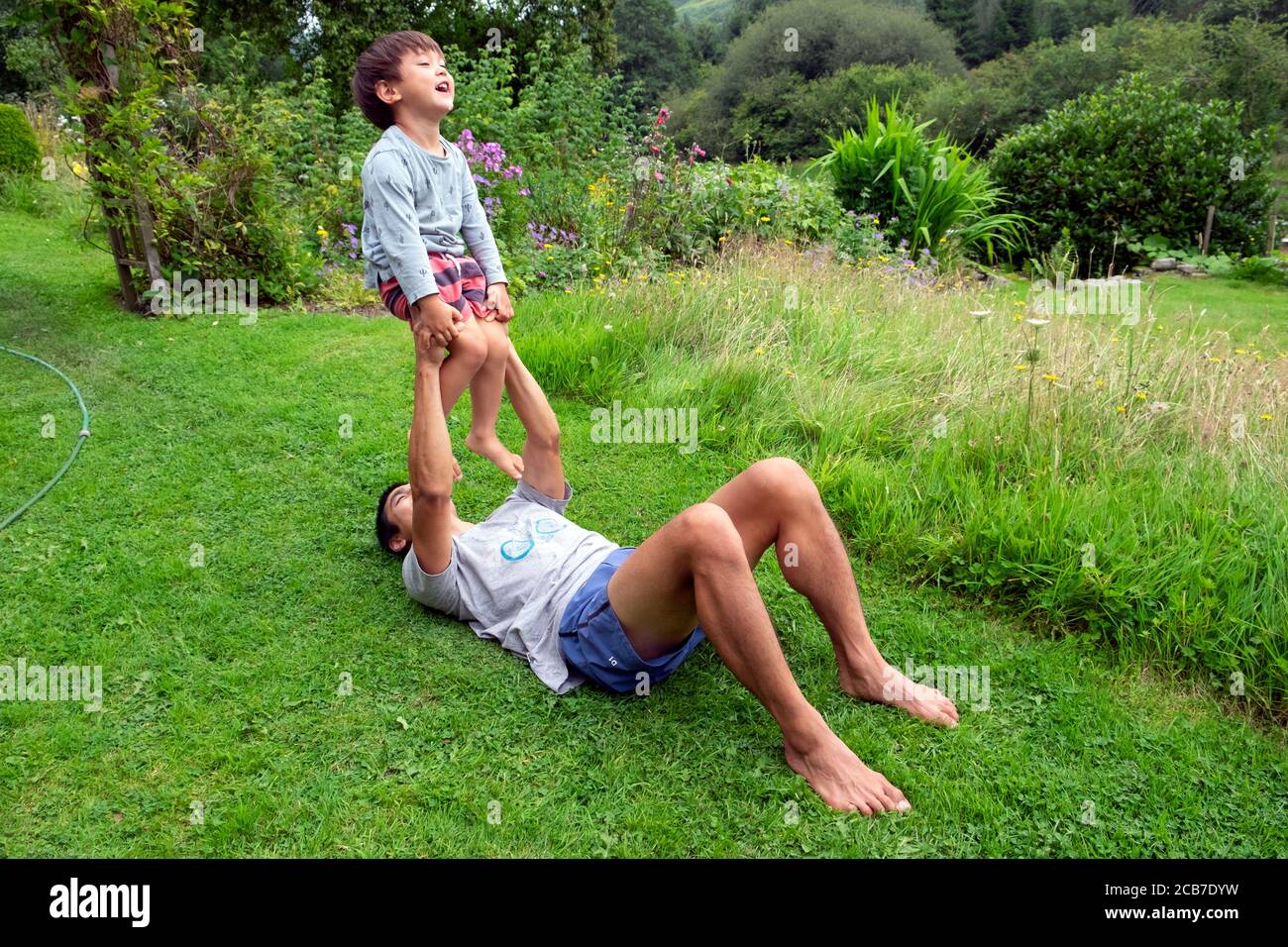 Father lying on the ground grass in country garden playing with balancing his 3 year old son child boy holding him in the air Wales UK KATHY DEWITT Stock Photo