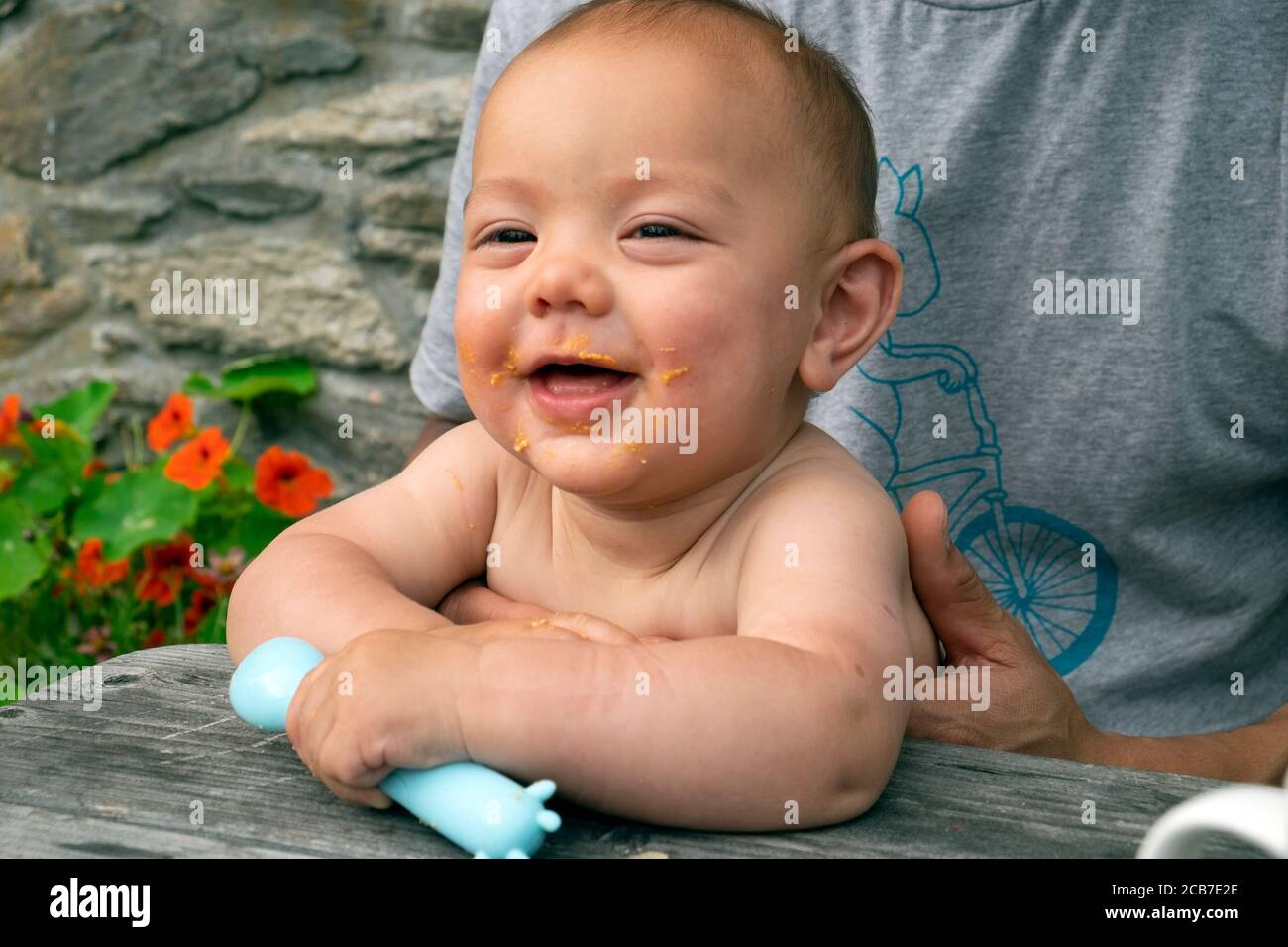 Mixed race smiling baby holding a plastic spoon sitting outside eating