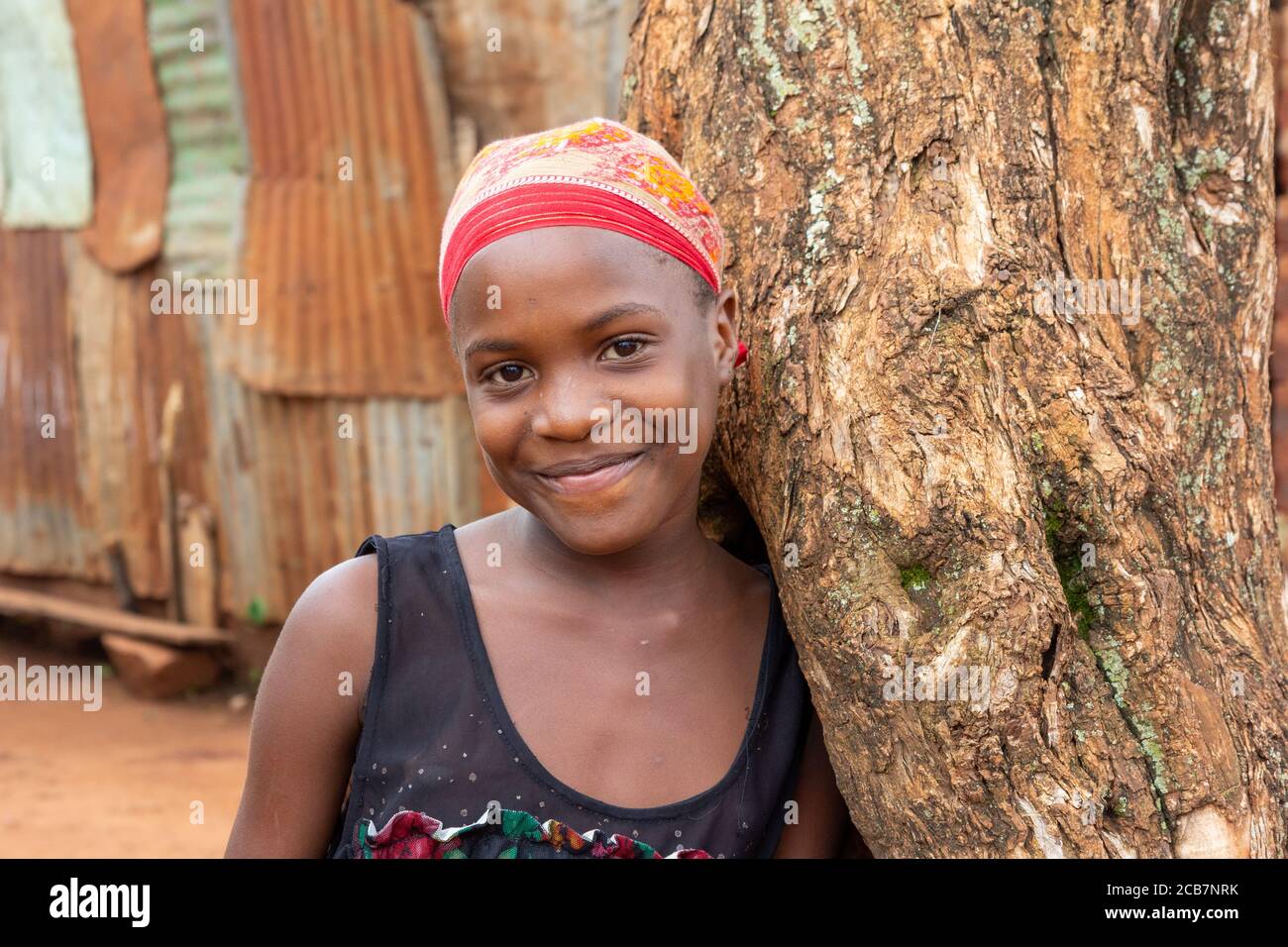 A beautiful teenage girl smiling and leaning against a tree. Shot in Uganda in 2017. Stock Photo
