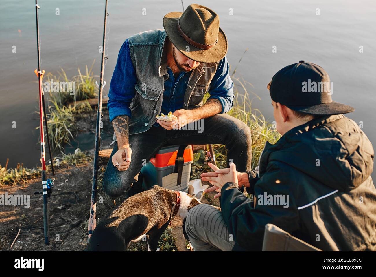 Caucasian adult father and teen son sit on fishing chairs and eat sandwich after good fishing. They tired, but happy Stock Photo