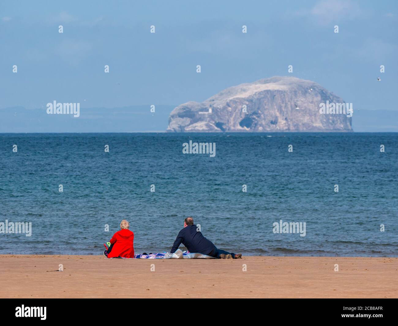 Couple sitting on beach admiring the sea view to Bass Rock gannet colony, Firth of Forth, Belhaven Bay, East Lothian, Scotland, UK Stock Photo