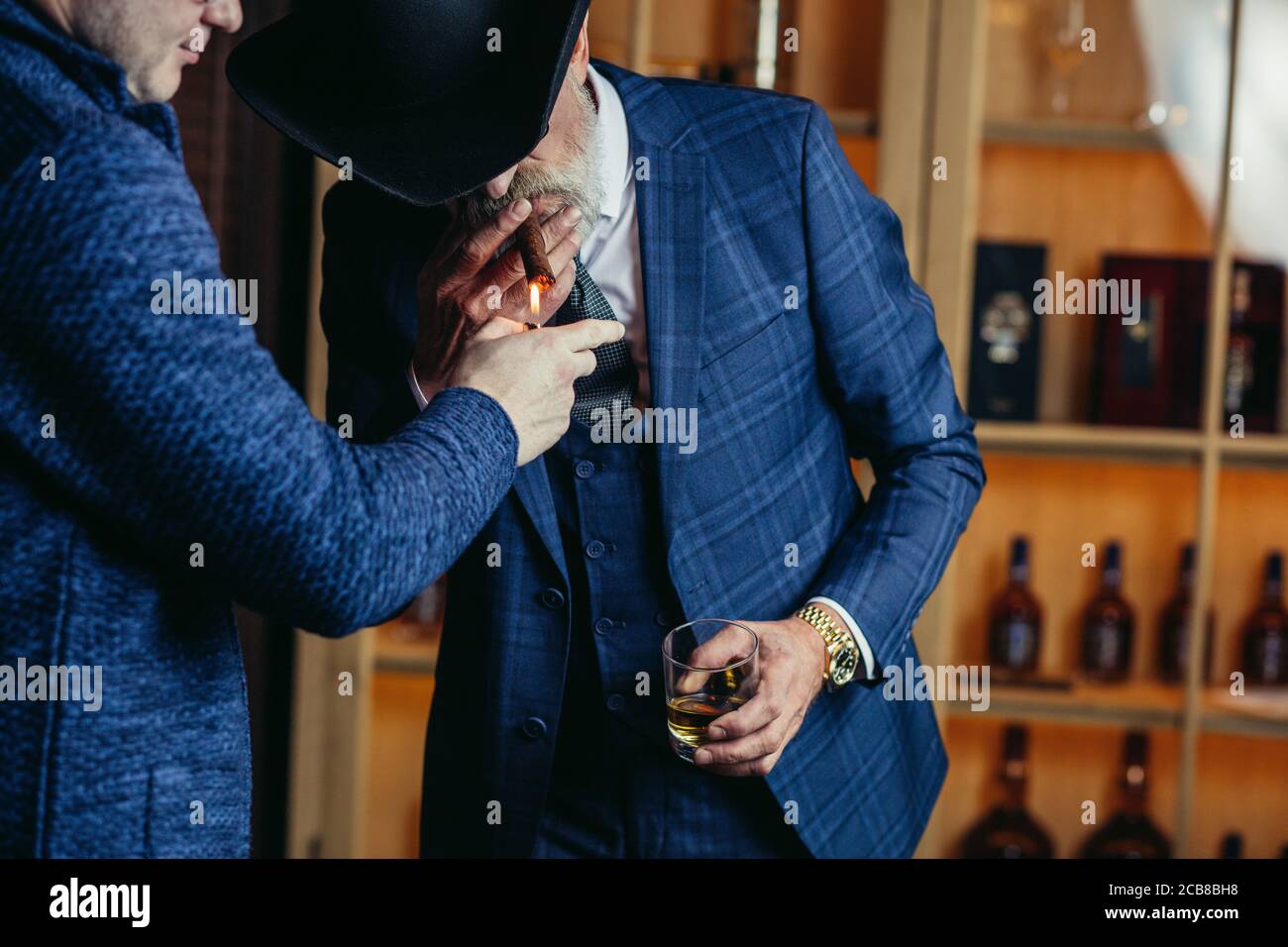 Elegant senior gentleman wearing spectacles and grey-haired beard standing at pub in wide-brimmed hat with glass of alcohol drink and cigar. Elderly m Stock Photo