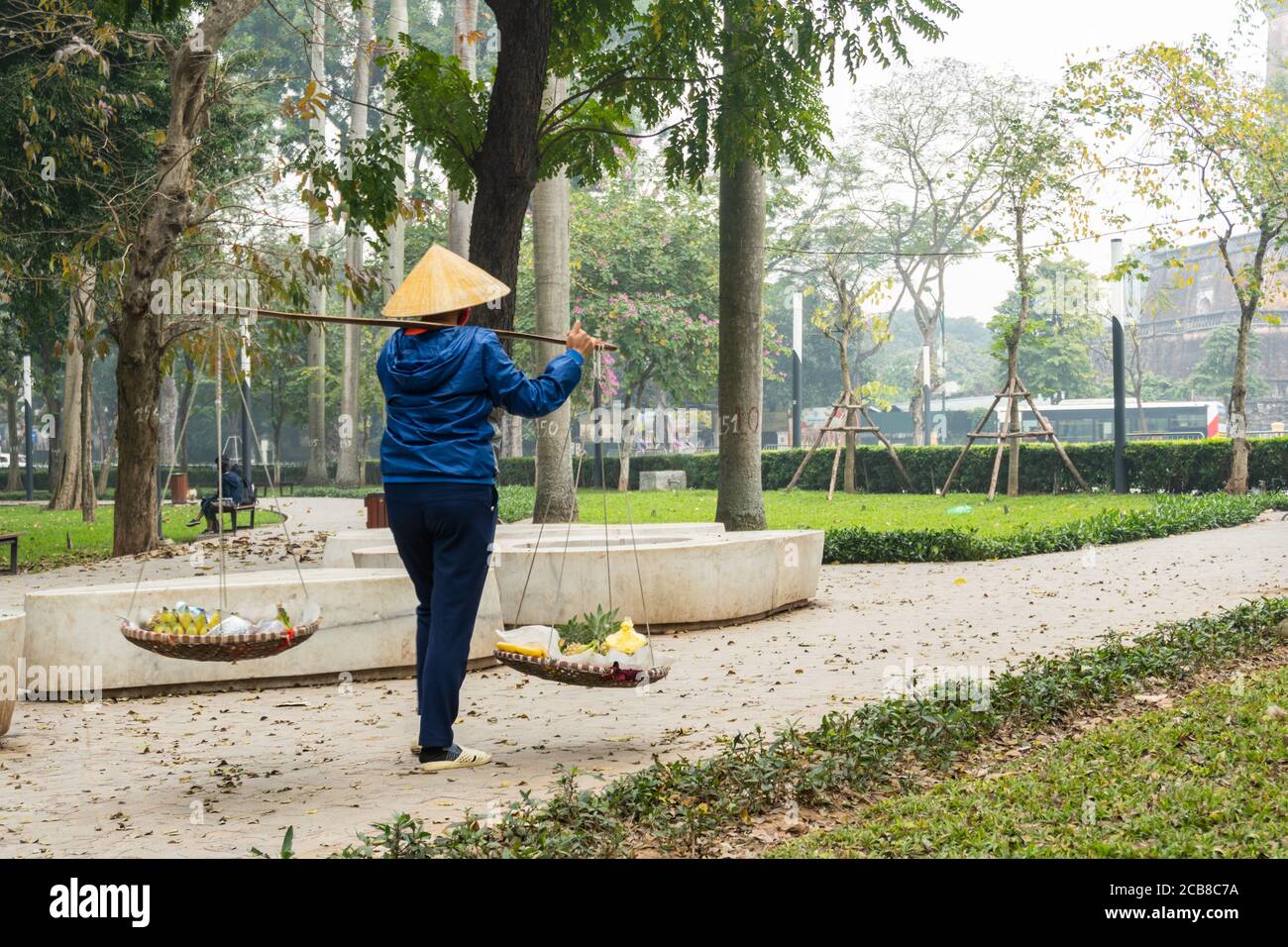 An elderly Vietnamese woman wearing a traditional cone hat walks with a traditional don ganh wicker basket yoke in a park. Street food vendor in Hanoi Stock Photo