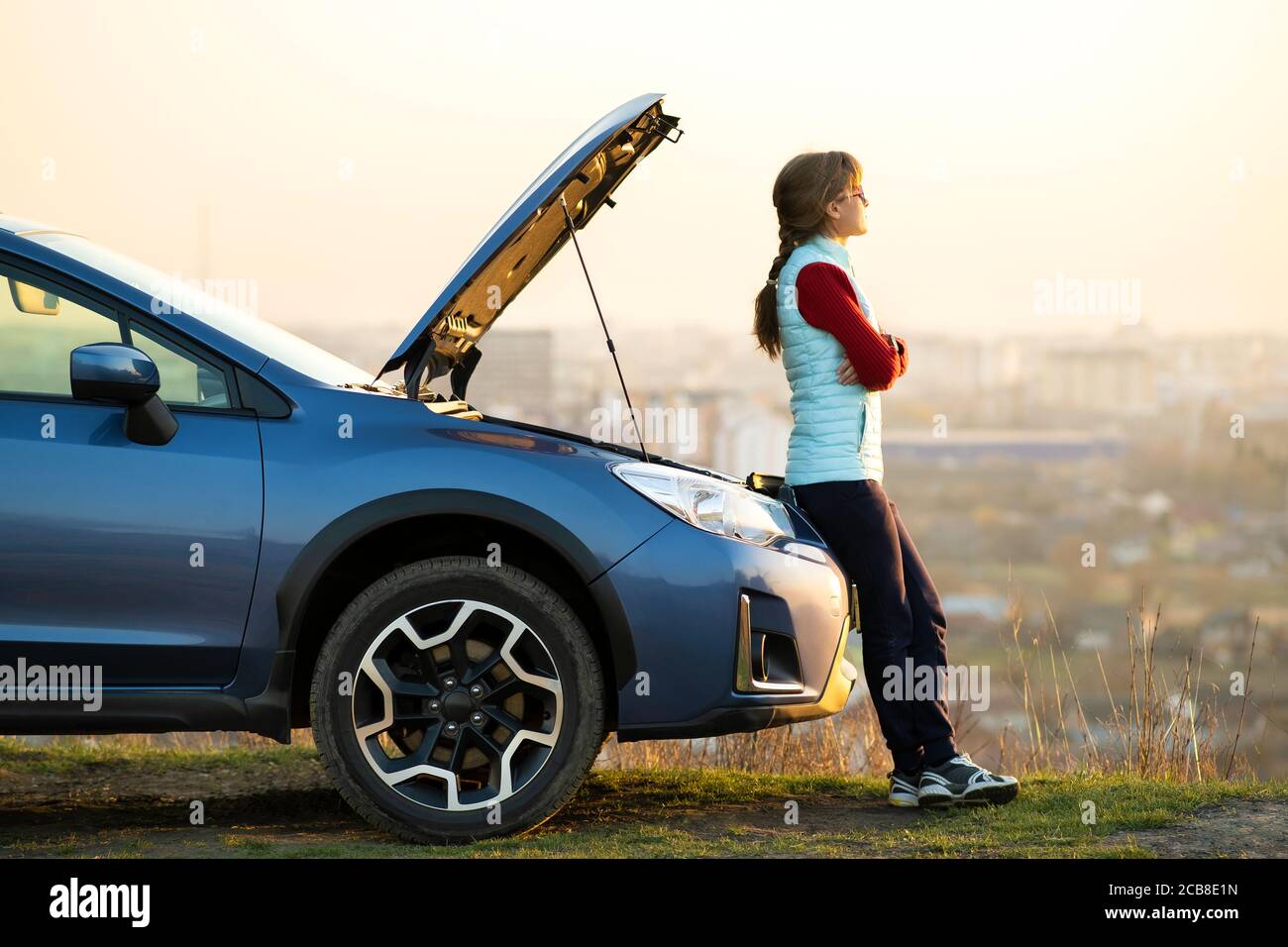 Young woman standing near broken down car with popped up hood having