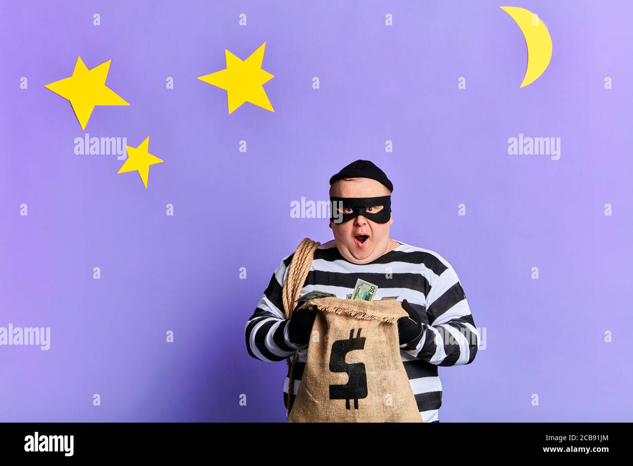 happy thief in mask and gloves with a rope on his shoulder opening a sack of money. isolated blue background. studio shot. night time Stock Photo