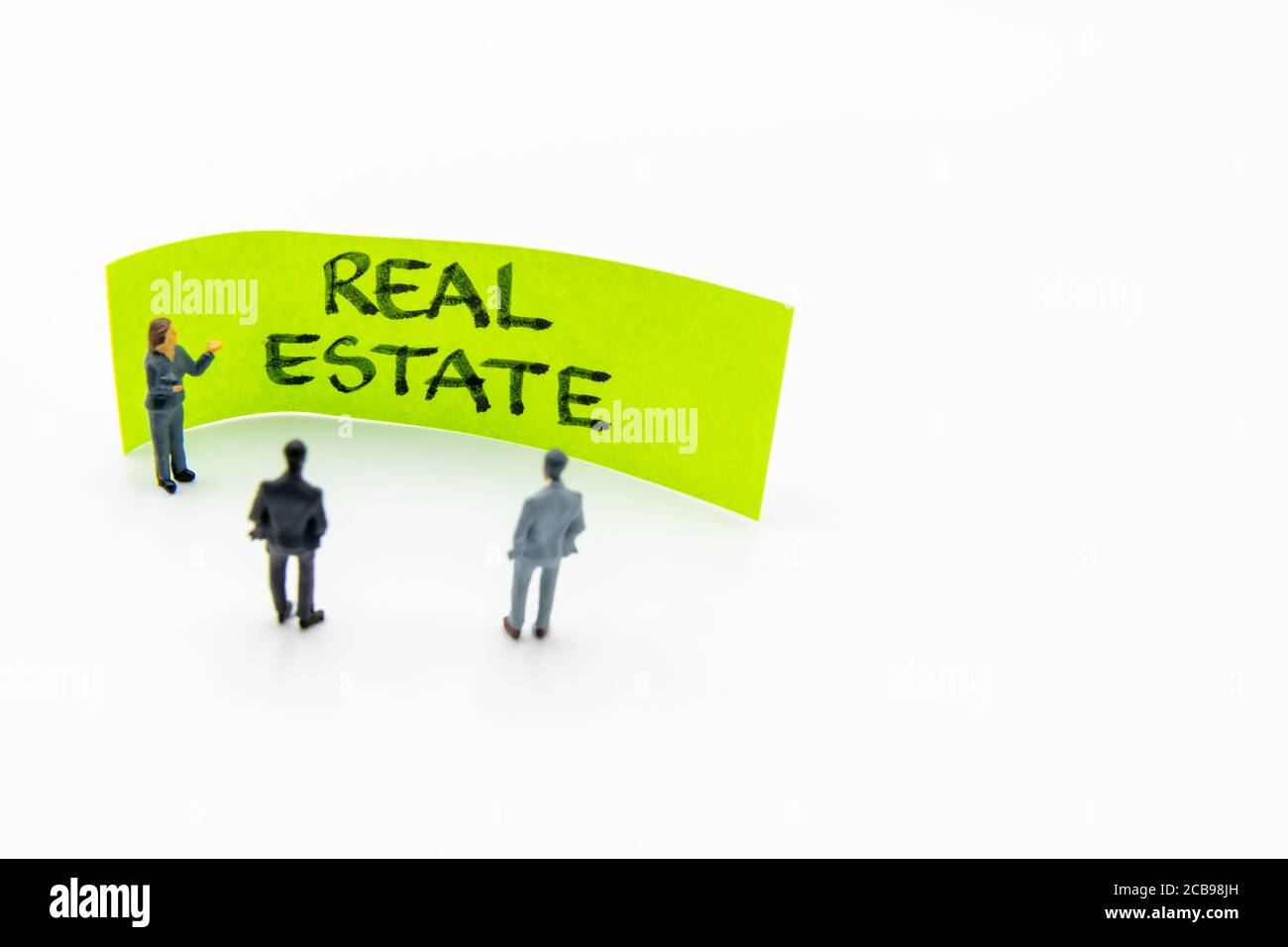 Presentation meeting with miniature figurines posed as business people standing in front of post-it note with Real Estate handwritten message in backg Stock Photo
