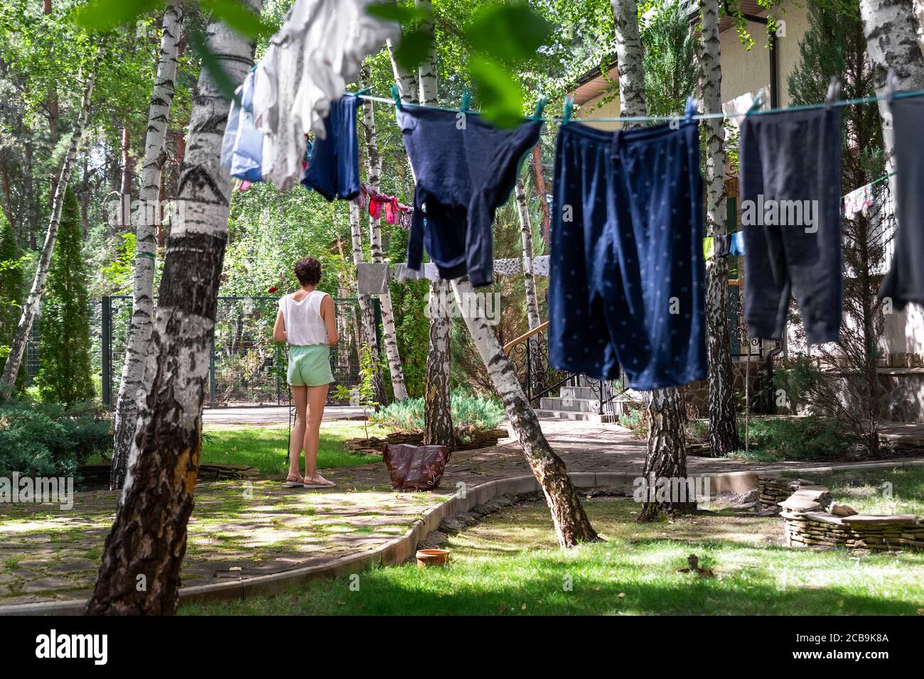 Candid real life portrait of young adult beautiful attractive caucasian woman hanging up fresh washed family clothes on birch tree clothesline with Stock Photo