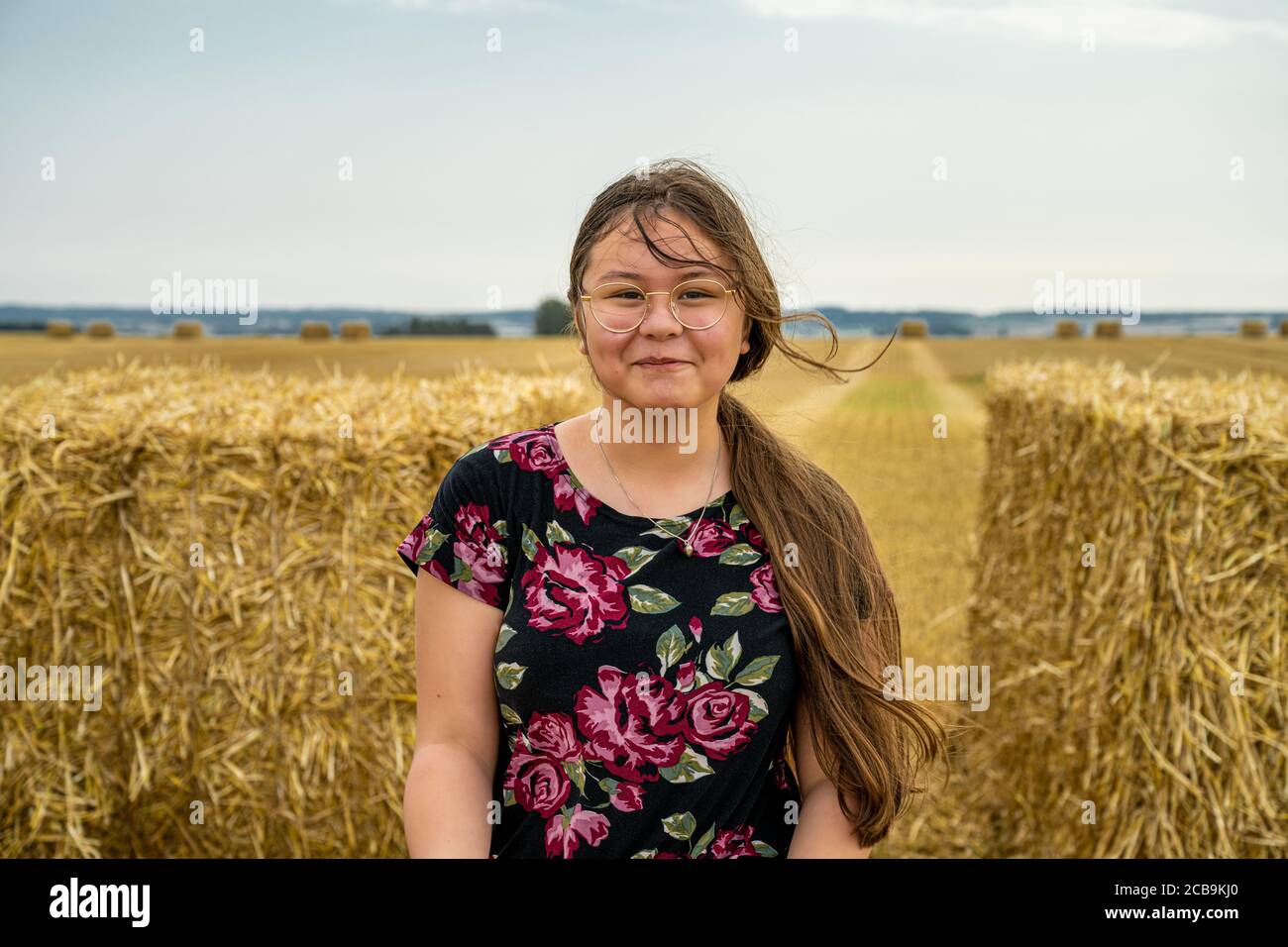 A preteen girl with long brown hair faces the camera and smiles. Bales of straw in a newly harvested field in the background Stock Photo