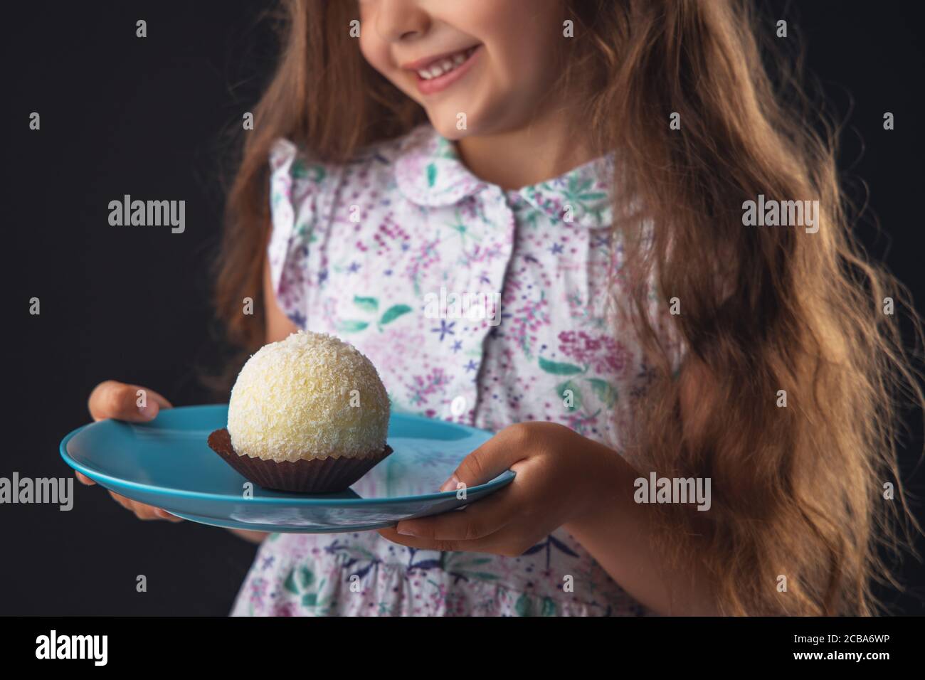 Beautiful little girl surprised from a tasty big chocolate coconut candy as a ball. Kid ready to eat cake. Stock Photo