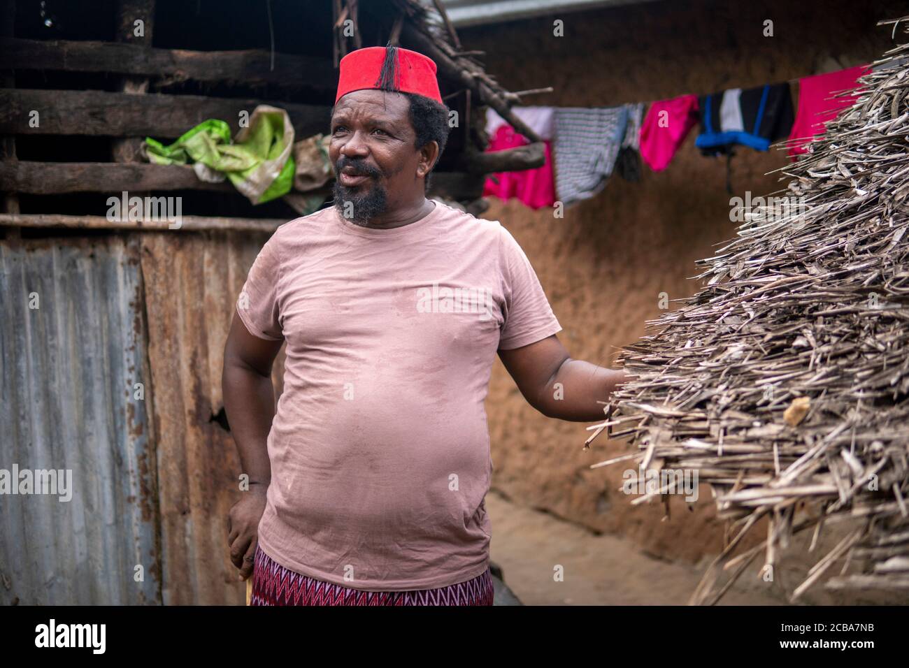 An African Older Man in Red Muslim Taqiyyah Fez Hat posing with a stick for lame people on Yard Near the Basic Hut with Thatched roof in Small Remote Stock Photo