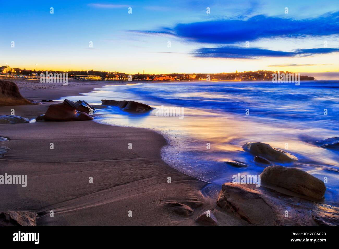 Sands and rocks of Bondi beach in Sydney at sunrise with blurred waves and waterfront of residential houses on North Bondi head. Stock Photo
