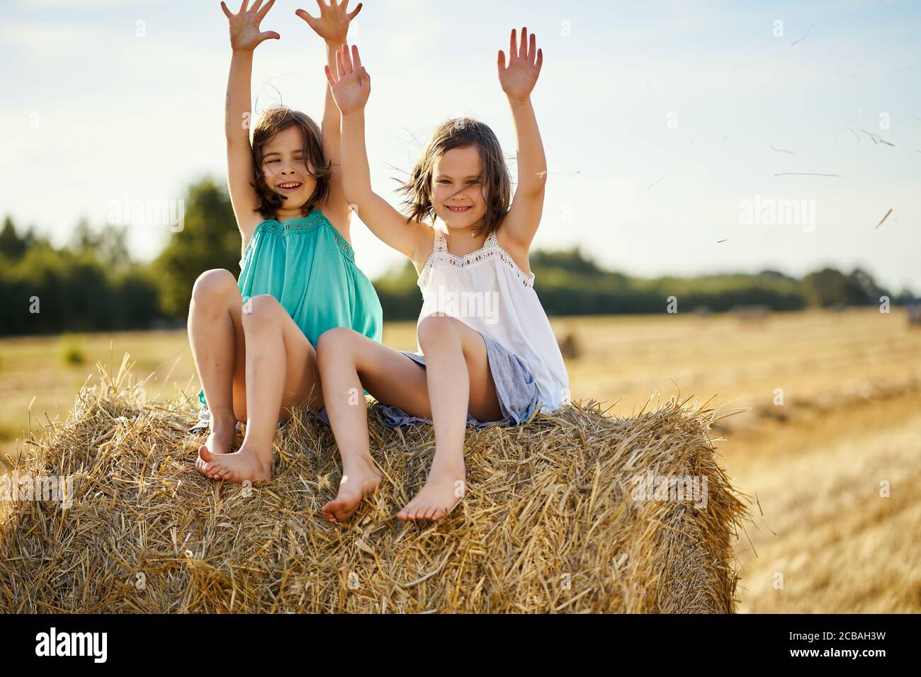 two charming girls are sitting on a roll of mown rye in a field Stock Photo
