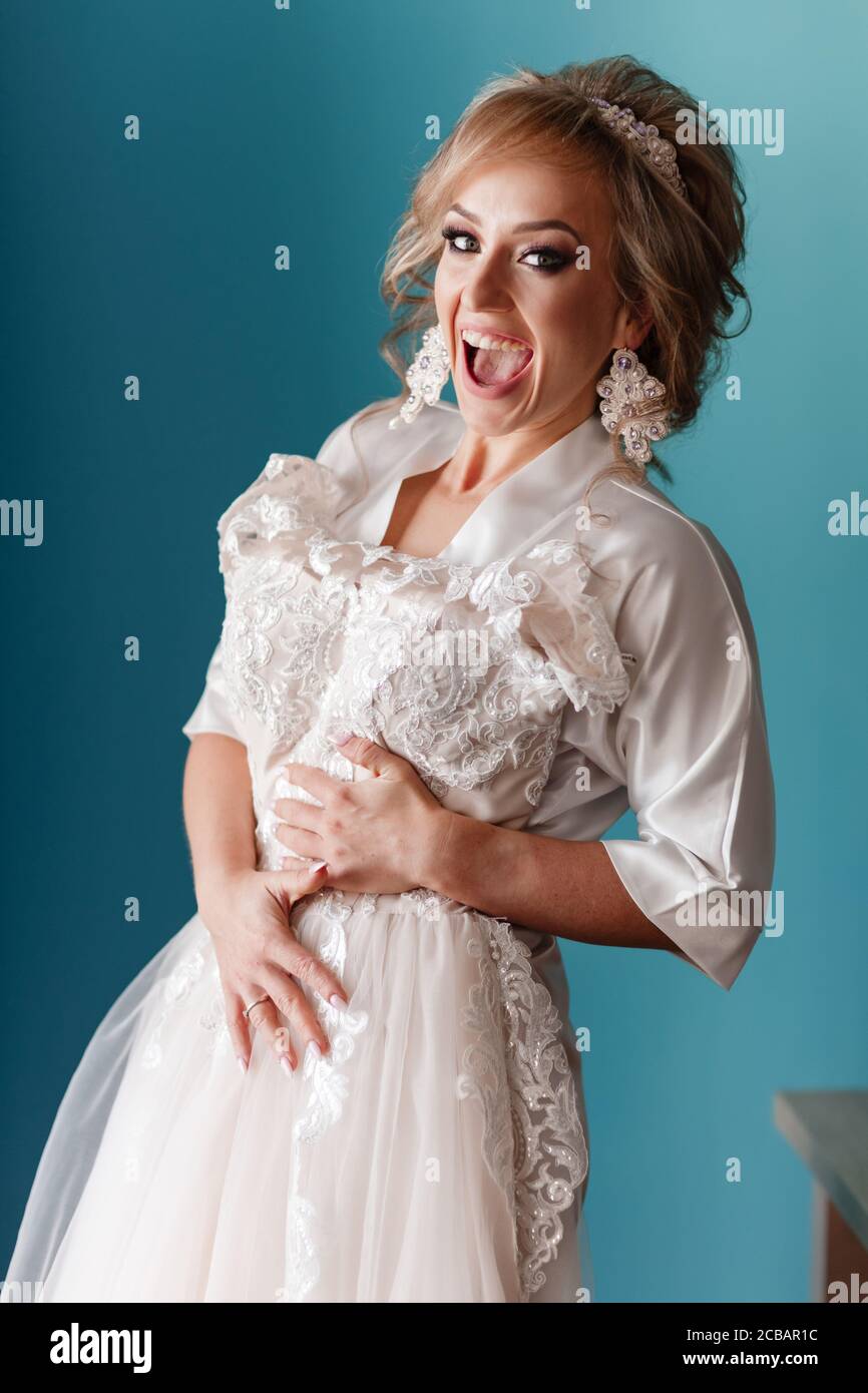 Bride morning preparation. happy beautiful young woman in silk bathrobe touching her wedding dress while standing on blue wall background Stock Photo