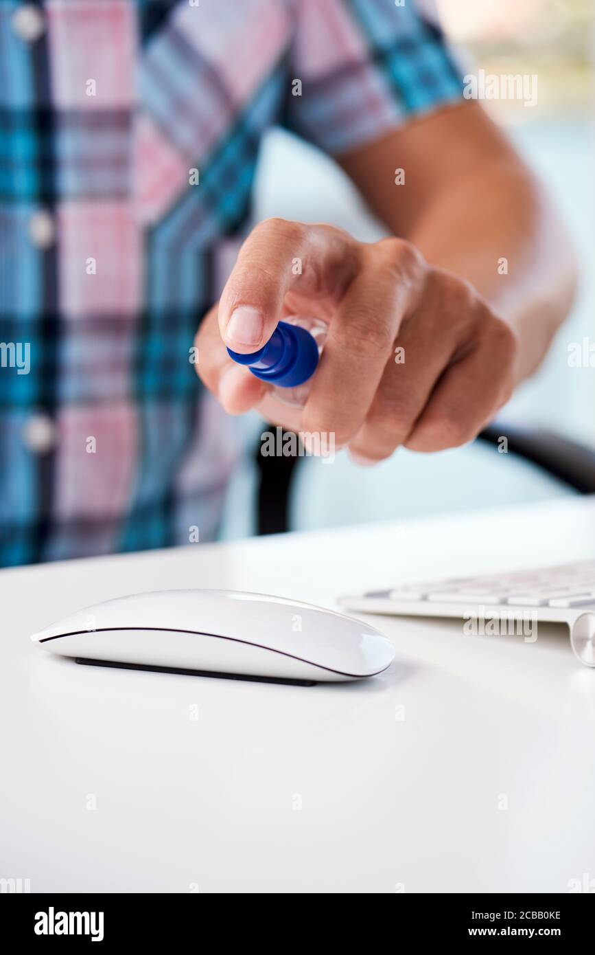 closeup of a young caucasian man sitting at his office desk disinfecting his computer mouse by spraying a disinfectant from a spray bottle Stock Photo