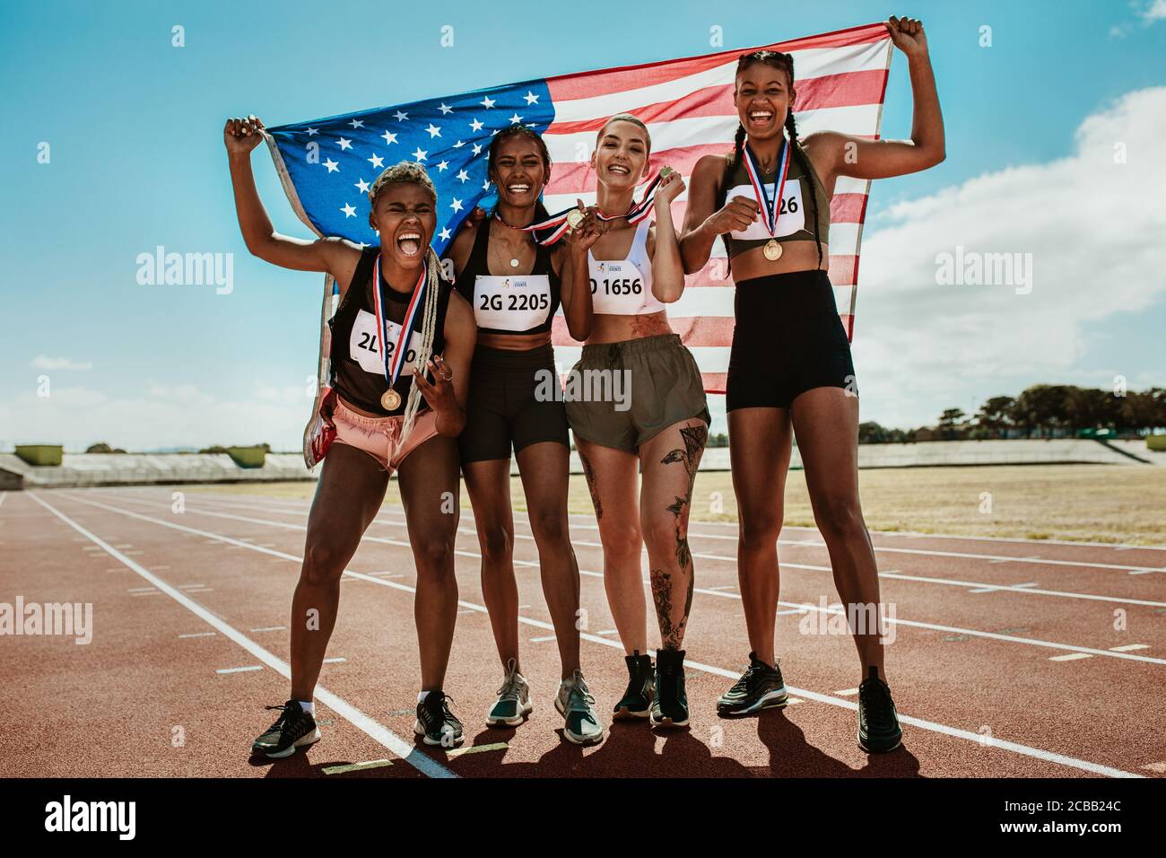 Happy multiracial athletes celebrating victory while standing together on racetrack. Group of runner with medals and American national flag winning a Stock Photo