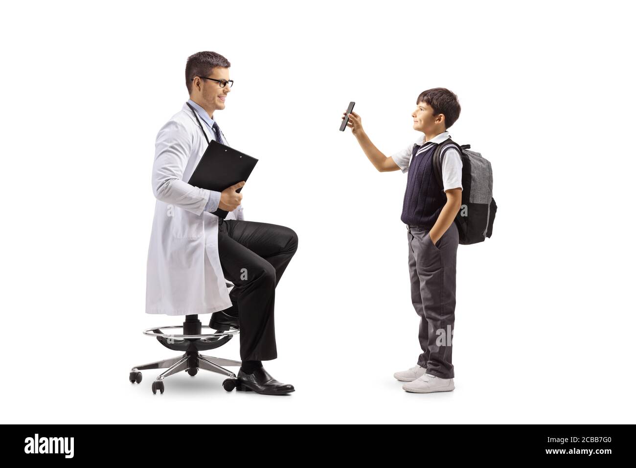 Full length profile shot of a schoolboy standing and showing a mobile phone to a male doctor seated on a chair isolated on white background Stock Photo