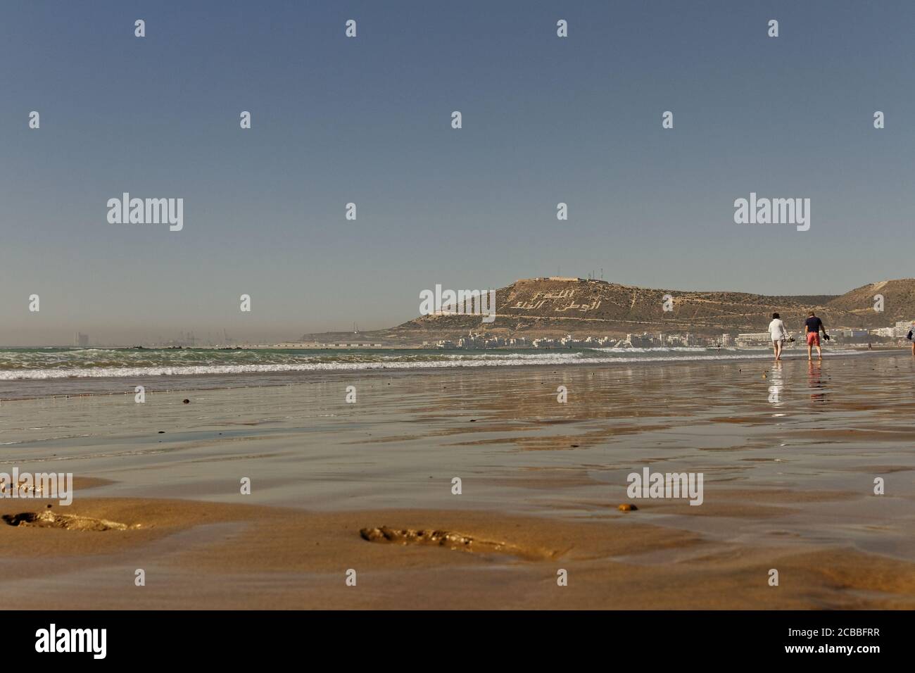 Agadir, Morocco.Feb.9,2019:Beach with tourist walking and Moroccan motto on the mountain. Writing on the hillside meaning, God, Country, King. People Stock Photo