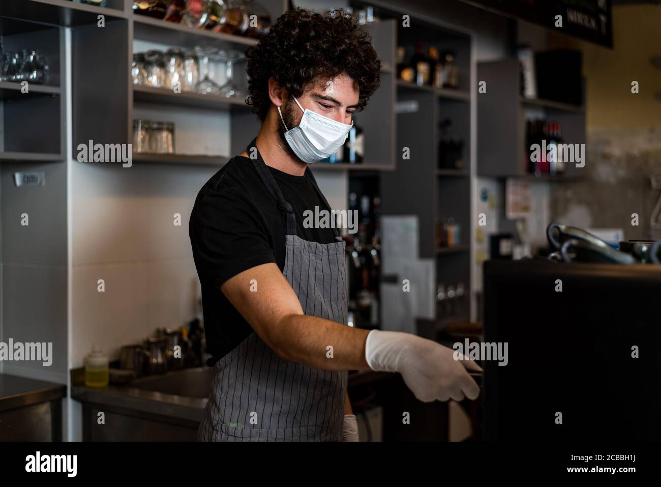 Young man, barista, making coffee espresso while wearing surgical mask and gloves for preventing corona virus spread - Bar safety working concept. Stock Photo