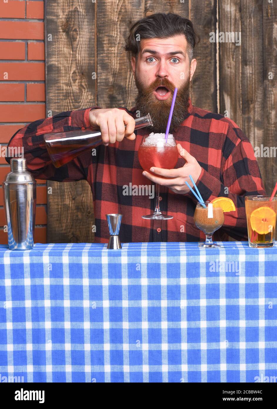 Barman with beard and surprised face makes cocktail, pouring glass with alcohol. Barman or hipster mixing drink. Drinks and party concept. Man holds wineglass and bottle on wooden texture background. Stock Photo