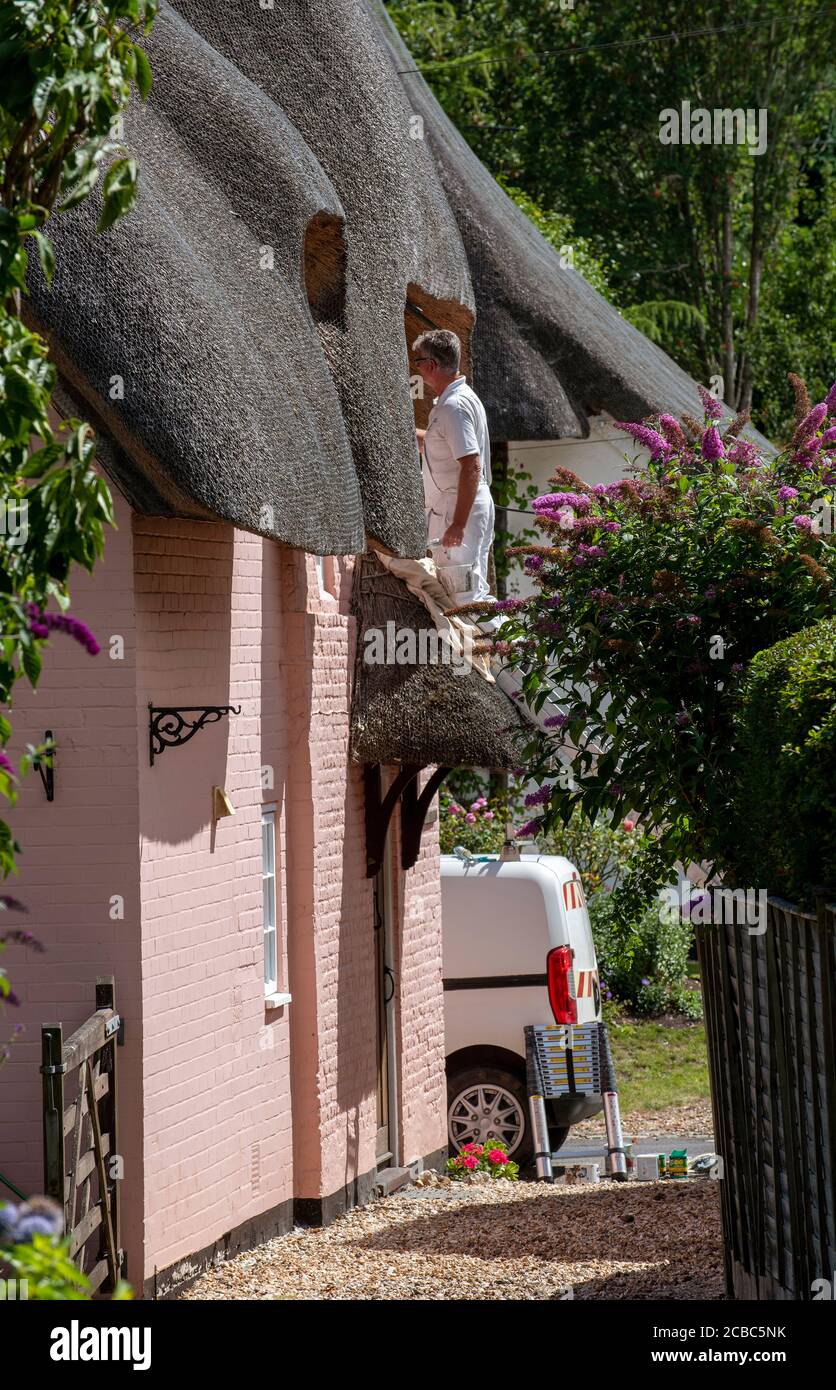 Hampshire, England, UK. 2020. Painter on a ladder using white paint to gloss the upper windows of an old thatched house in the UK Stock Photo