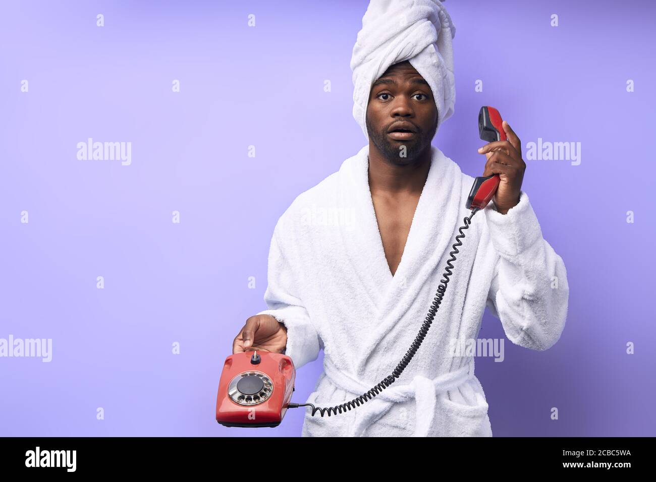 Surprised african man in bathrobe and towel with opened mouth. Using landline phone for talking with friend, retro style. Isolated over purple backgro Stock Photo