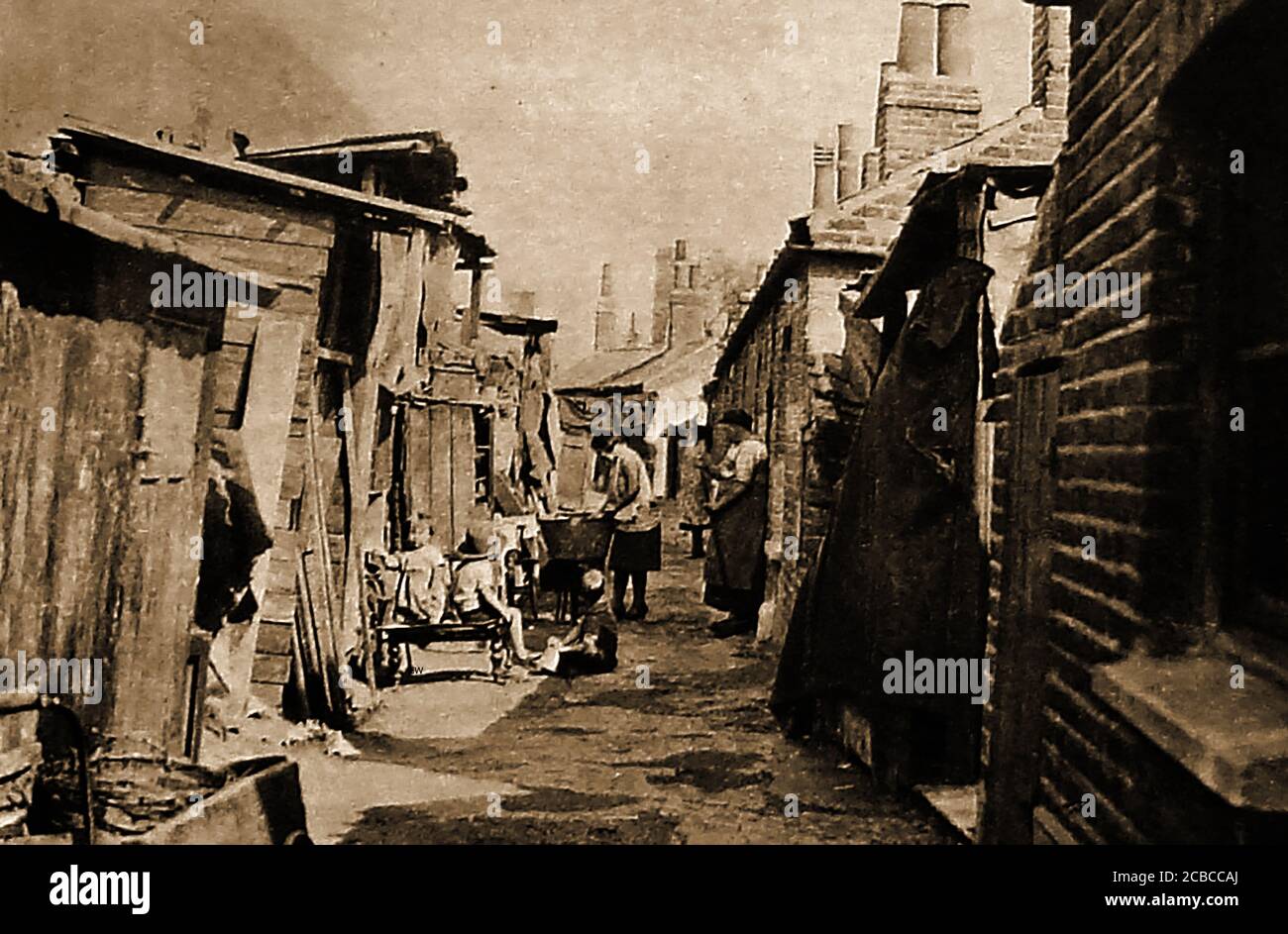 C 1940's -A back street scene between two rows of Britain's back to back houses & slums. Children are seen playing whilst a woman washes he clothing in an enamel bath tub. Lean-to wooden sheds have been roughly constructed behind some dwellings. Stock Photo